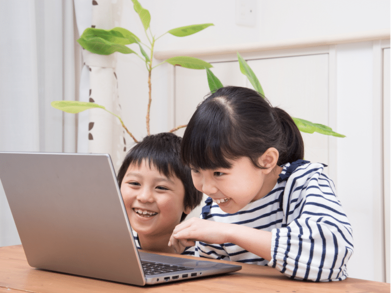 A wider shot showing two children sitting together at a table, both laughing as they look at a laptop in front of them. The children are dressed in striped shirts, and a plant can be seen in the background. This is the full version of the close-up shown above.