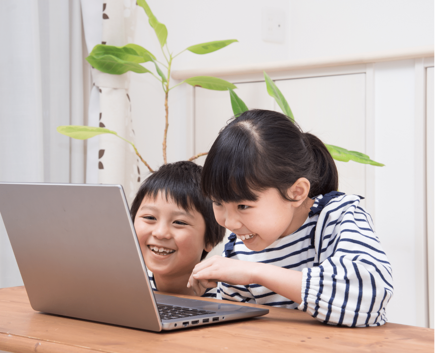 A wider shot showing two children sitting together at a table, both laughing as they look at a laptop in front of them. The children are dressed in striped shirts, and a plant can be seen in the background. This is the full version of the close-up shown above.