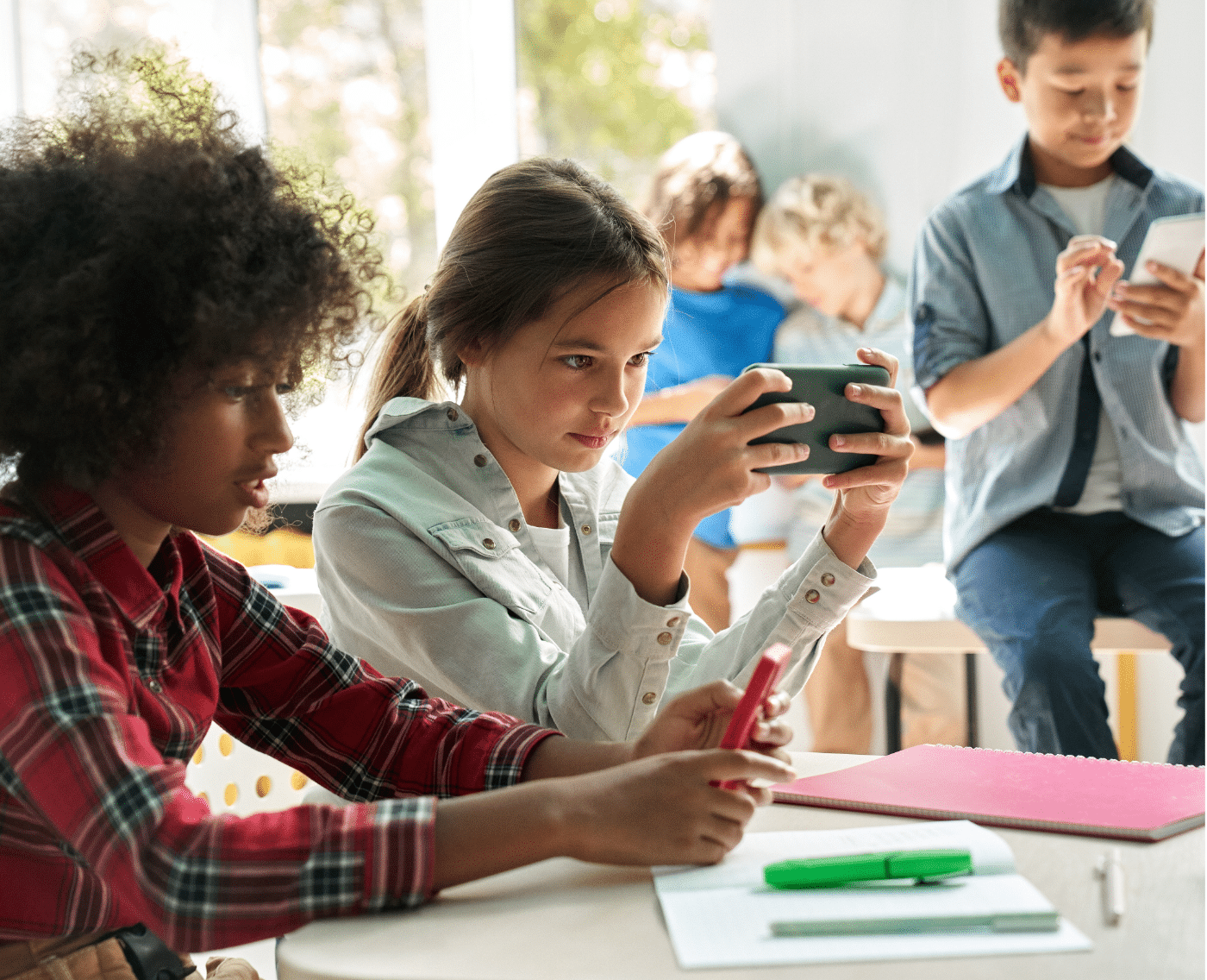 A classroom setting with a group of children engaging with digital technology. Two girls sit at a table in the foreground; one in a plaid shirt is holding a smartphone with a red cover, while the other in a white jacket is focused on her own smartphone. The table has school supplies, including a notebook and markers. In the background, three boys are sitting on a desk, interacting with tablets and smartphones. The scene is well-lit, with natural light streaming through large windows.