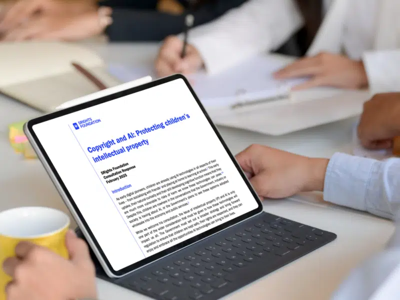 A group of professional people sat at a table. The image is framed to only see four people's sets of hands and the desk. The focus is on a propped-up tablet that is displaying 5Rights' consultation response, a non-descript document with the 5Rights logo and an electric blue title.