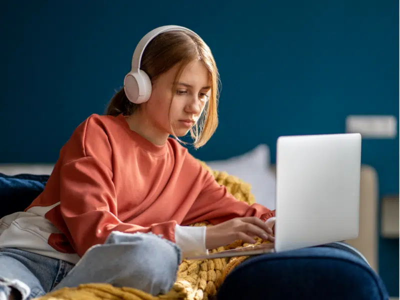 A wider shot of the same teenage girl seated comfortably on a couch at home. She is wearing a rust-orange sweatshirt, light blue ripped jeans, and white over-ear headphones, focused on her laptop. A yellow knitted blanket and blue wall add to the cozy setting. This image is a fuller version of the close-up shown above on the same page.