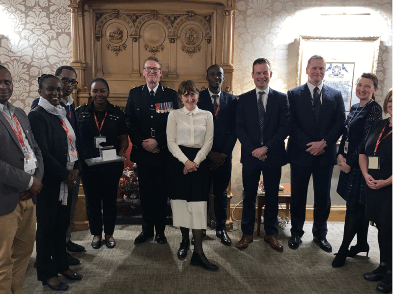 A group of people, including Rwandan Ministry representatives, Scottish Police officials, and Baroness Beeban Kidron (woman in the centre wearing a white shirt), stand together in a formal setting with an ornate wooden fireplace in the background. The group is smiling and dressed in formal attire.